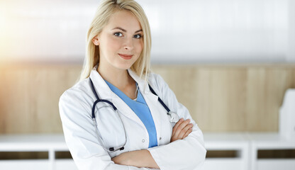 Woman-doctor at work while standing straight in sunny clinic. Blonde cheerful physician ready to help patients