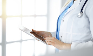 Unknown woman-doctor standing in sunny clinic and using tablet pc, close-up. Data and best service in medicine