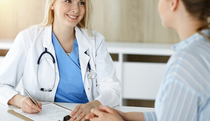 Fototapeta premium Woman-doctor and patient sitting and talking in sunny clinic. Blonde therapist is cheerfully smiling