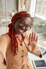 High angle portrait of young African-American woman waving at camera while taking selfie photo or...