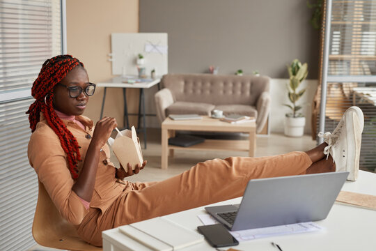 Full Length Portrait Of Young African-American Woman Eating Takeout Food And Looking At Laptop Screen While Relaxing At Home Office, Copy Space