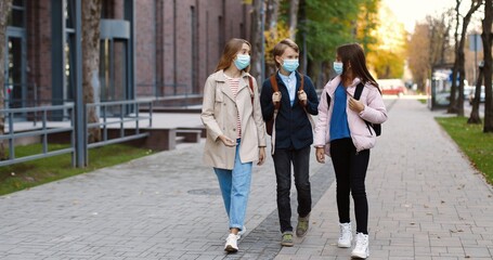 Cheerful Caucasian teen children walking after school and talking in good mood. Boy speaking with beautiful happy girls in masks. Male and female pupils and friends on street. Classmates concept