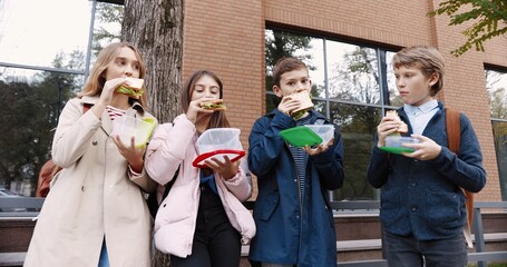 Portrait of joyful Caucasian young school students standing outdoor with food containers and eating sandwiches. Boys and girls classmates with backpacks having snack on lunch near school on break.