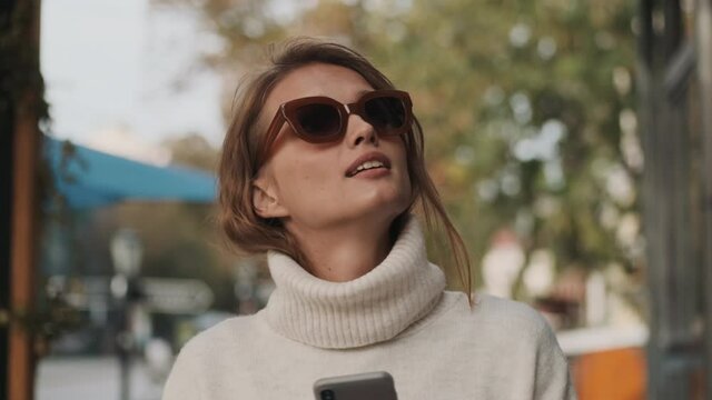 Pretty fashion girl in sunglasses dressed in white cozy sweater chatting with friends on smartphone dreamy walking through city street