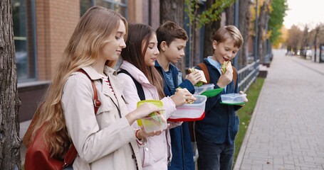Portrait of happy Caucasian school teenagers standing outdoor with food containers and smiling while eating sandwiches. Joyful boys and girls pupils with backpacks having snack near school on break