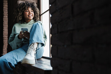 African american woman using mobile phone while sitting on windowsill