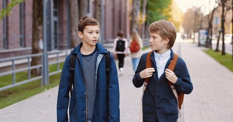 Caucasian happy nice male school students walking on street after school and chatting. Portrait of joyful young boys pupils with backpacks in good mood speaking outdoors in city. Classmates concept