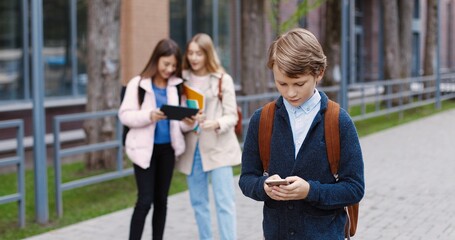 Fototapeta premium Portrait of Caucasian school student boy going to school and typing on cellphone outdoors. Male pupil texting and tapping on smartphone. Girls tapping on tablet on background. Pupils concept
