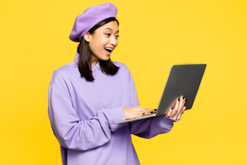 happy asian woman in beret using laptop isolated on yellow