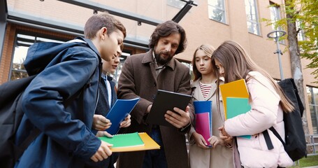 Portrait of Caucasian children with colorful copybooks standing around man educator near school on...