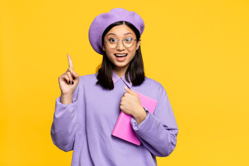 excited asian woman in beret and eyeglasses holding notebook and pointing with finger isolated on yellow
