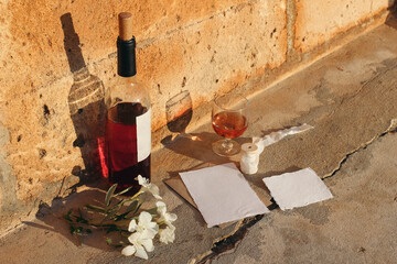 Mediterranean summer wedding composition. Blank paper greeting card mockups with red wine bottle, drinking glass and white oleander flowers. Orange sandstone grunge ground in sunrise sunlight.