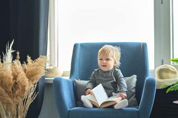 Infant child baby girl toddler sitting and reading book on the sofa in the living room. portrait