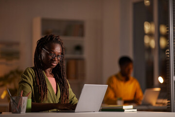 Portrait of young African-American woman wearing glasses while using laptop in office at night, copy space