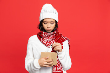 sad asian woman in hat and scarf holding tissue box isolated on red