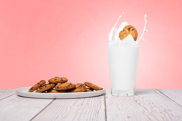 Chocolate chip cookies splashing falling into a full glass of milk with cookies on the side sitting on a wood surface against a pink texture wall with copy space and room for text on a horizontal imag