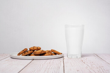 Chocolate chip cookies sitting next a full glass of milk on a wood surface against a textured wall background with copy space and room for text on a horizontal image