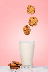 Chocolate chip cookies splashing falling into a full glass of milk with cookies on the side sitting on a wood surface against a pink texture wall with copy space and room for text on a vertical image