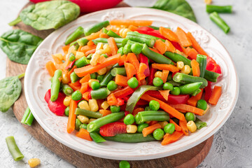 Various steamed vegetables close-up. Boiled peas, corn, string beans, carrots, and peppers in a white plate on a gray concrete table. Diet and vegan food.