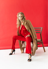 A beautiful blonde curly girl wearing a red pantsuit, unbuttoned on her braless big breasts and coat, sits on a chair on a red background. Advertising, fashionable, commercial design