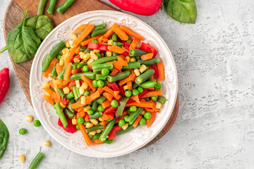 Various steamed vegetables top view. Boiled peas, corn, string beans, carrots, and peppers in a white plate on a gray concrete table. Copy space for text. Diet and vegan food.