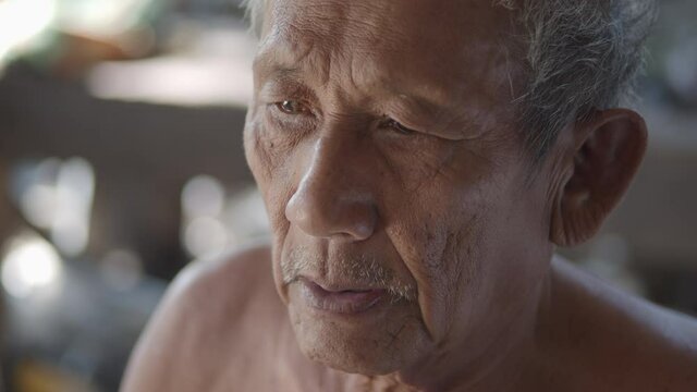 Slow motion of white-haired Asian elderly man do not wear clothes because of hot weather, poor status, more than seventy is sitting smoking tobacco wrapped in nypa fruticans (nipa palm)leaves.