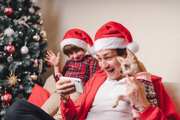 Father with child and puppy dog in Santa hats having a video chat on Christmas holidays at smartphone, sitting on a couch in the living room with Christmas tree at home.