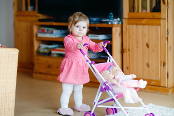 Cute adorable baby girl make first steps and push with doll carriage. Beautiful toddler child pushing stroller with toy at home. Happy kid with two dolls