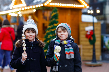 Little cute kid girl and boy eating white chocolate covered strawberries and apple on skewer on traditional German Christmas market. Happy children, best friends, twins and siblings on snowy day