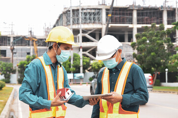 Construction engineer wearing a helmet uses a tablet while standing outdoors at a construction site.