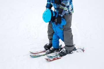 Tired toddler boy in warm blue overalls learns to ski with his dad during winter break in snowy mountains. cold day, useful activities with parents. Ski training. Sports education, care, support