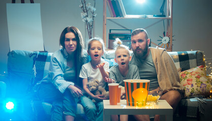 Expressive. Happy family watching projector, TV, movies with popcorn and drinks in the evening at...