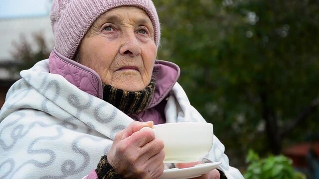 Old grandmother with a cup of tea in cold weather. An old woman is drinking hot tea, covered with a blanket. An old grandmother walks with tea in cold weather. The concept of comfort and family care