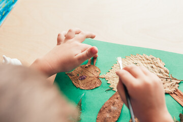 child glues an applique made of autumn leaves, view from back, close up