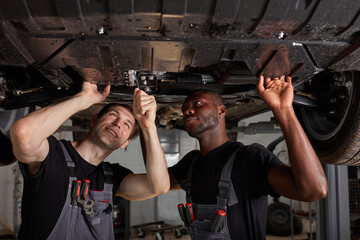 two interracial diverse mechanics checking car bottom in auto service, cooperate, work together