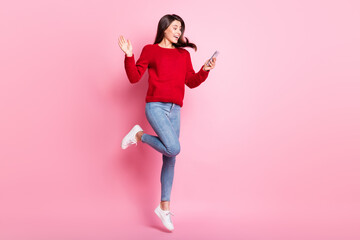 Full length photo of pretty girl waving hand screen hold cellphone jump wear red pullover jeans sneakers isolated pink color background