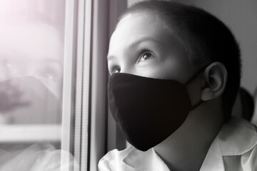 A little boy in a black mask at the window, self-isolation and quarantine, coronavirus.