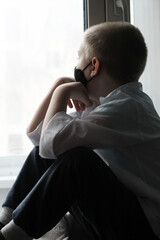 A little boy in a black mask at the window, self-isolation and quarantine, coronavirus.