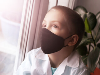 A little boy in a black mask at the window, coronavirus.