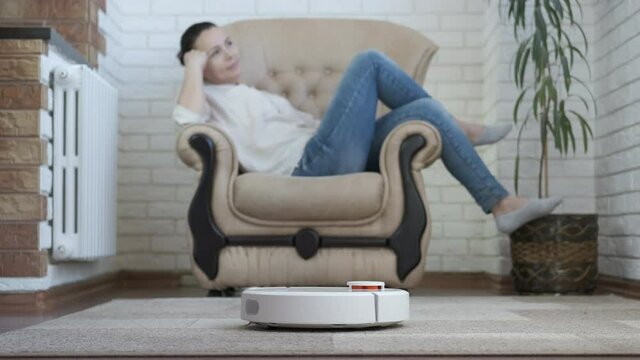 Relaxing with a robot vacuum cleaner. The female is resting in the chair, the smart vacuum cleaner is working.
