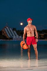 Handsome lifeguard man with a naked torso with a buoy in his hand posing on the beach at sunset