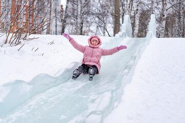 A young woman in a pink down jacket is riding in a city park on an ice slide without a sled