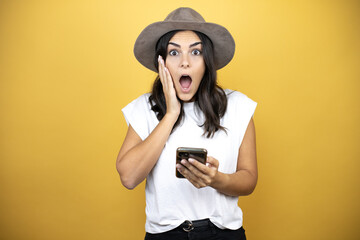 Beautiful woman wearing casual white t-shirt and a hat standing over yellow background chatting, surprised and looking her phone