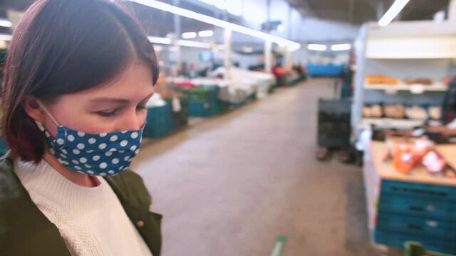 A woman in a mask walks through a vegetable market looking for a food item to buy. Shopping at retail, realisation of consumerism.