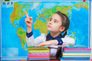 Portrait of a smart cute girl hugging a lot of books on the background of the world map. Points finger, gives sign