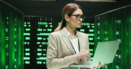 Caucasian young woman in glasses standing at servers with laptop computer, typing on keyboard and checking big data processor. Female analytic in data storage working on digital information security.