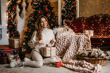 Young smiling woman picks a box with a Christmas present in her hands, relaxing at home, enjoying Christmas. A beautiful girl at home, sitting near the Christmas tree. Gift for the new year