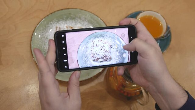 Taking picture with smartphone of a tasty dessert and sea buckthorn tea. Slice of Napoleon cream cake, made from puff pastry, lies on a plate on a table in a restaurant or cafe.