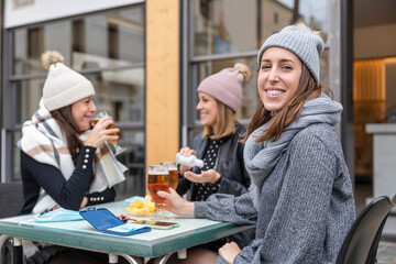 Young smiling women wearing winter clothes drinking beer on a bar. One woman is disinfecting her hands with gel. One woman is looking at camera. New normal concept
