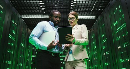 Mixed-races man and woman with tablet device discussing issue at datacenter. Multiethnic developers standing at servers storage and looking at gadget. Male and female analytics colleagues talking.